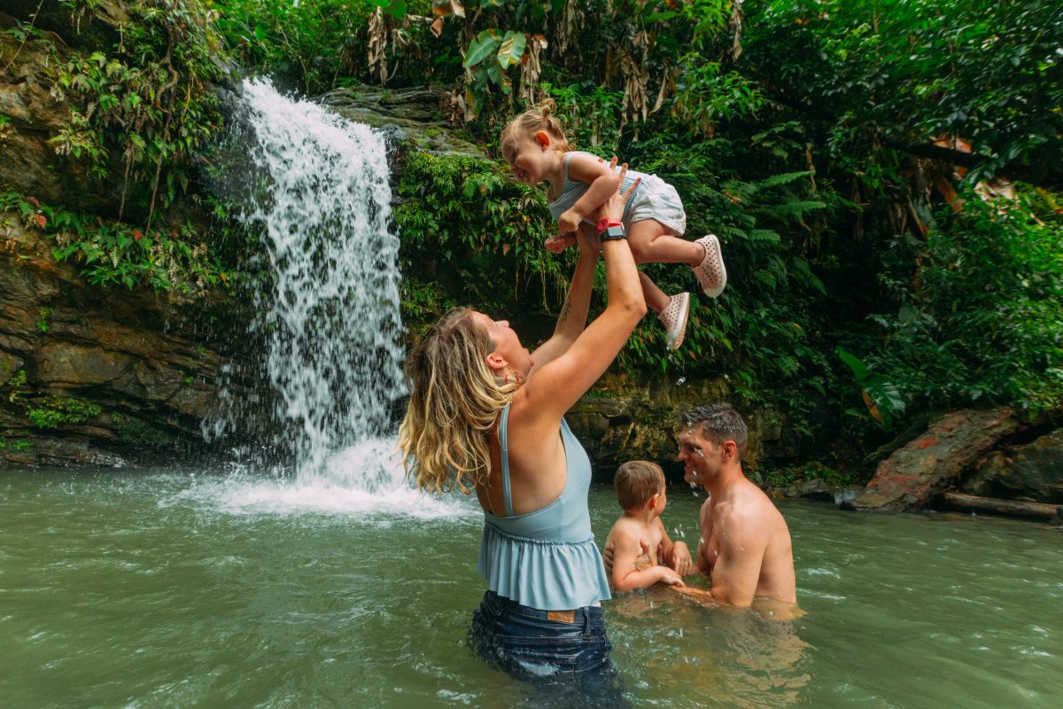 Mother holds a baby in the air while being half submerged in a forest waterfall pool.