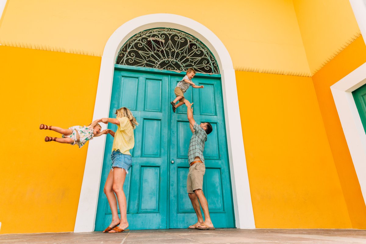 A couple plays with their children. The family stands next to a vivid yellow wall with a teal blue door.
