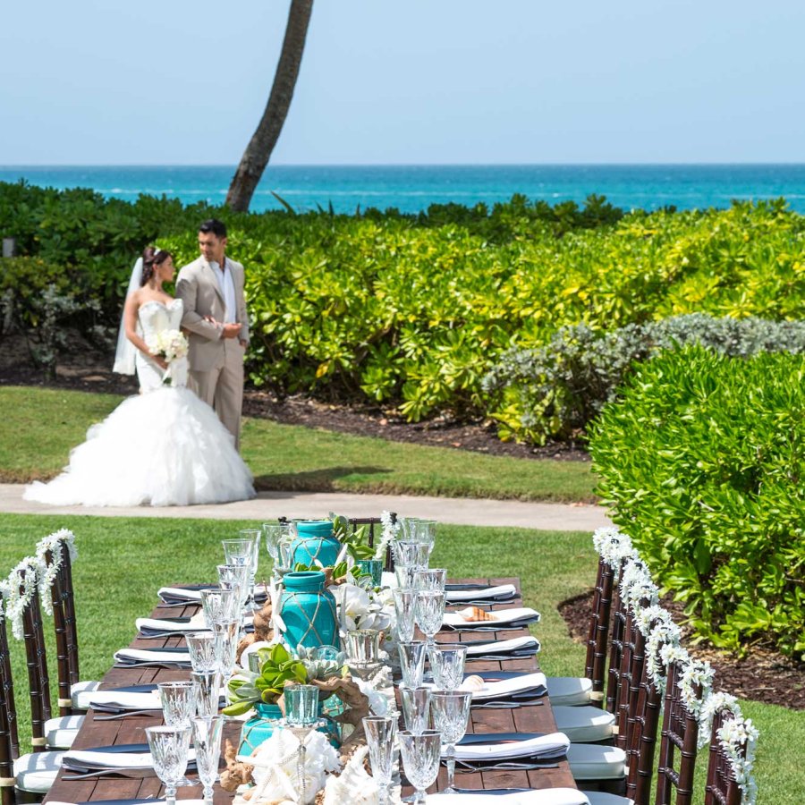 A couple in wedding garb walks down a pathway surrounded by greenery, with the ocean behind them.