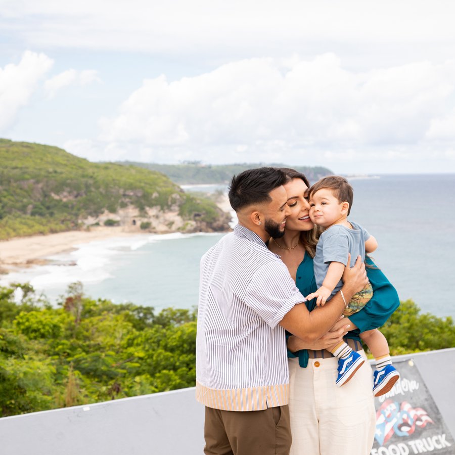 A couple holds a baby close to them, smiling. They stand in front of a coastline.