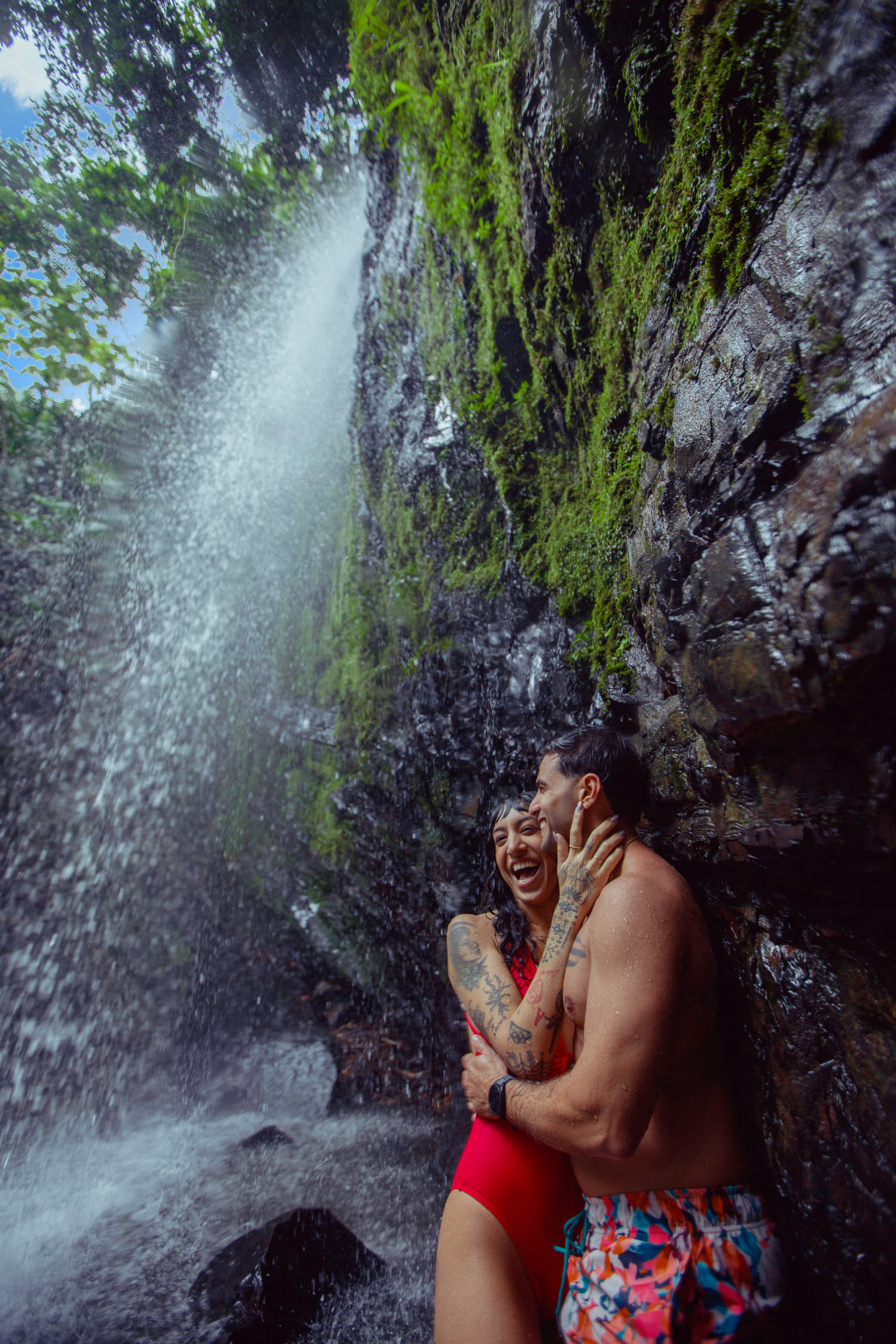 A woman and a man in bathing suits hold each other while laughing as they stand underneath a mossy cliff with a waterfall sprinkling off of it.