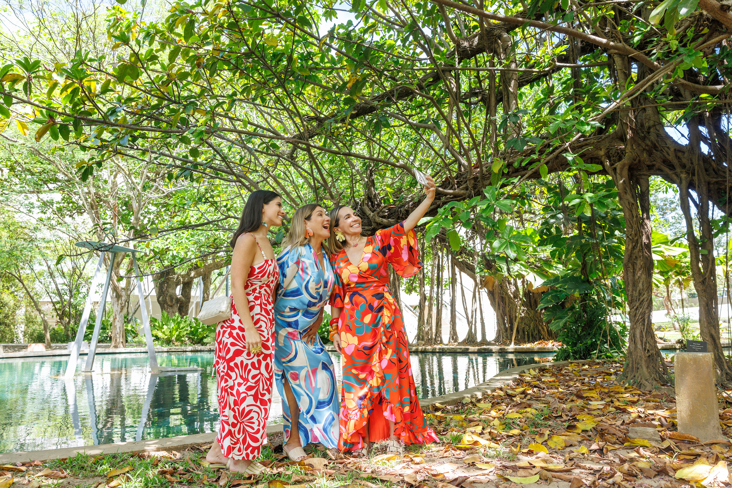 Three visitors taking a selfie beside a reflective pool beneath sprawling tropical trees and hanging roots in a lush garden setting in Puerto Rico.
