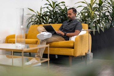 A man works on his laptop in a modern office while sitting on a yellow sofa.