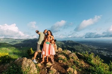 A mother, father, and two young girls pose for a selfie on top of a mountain with a dramatic backdrop in Puerto Rico.