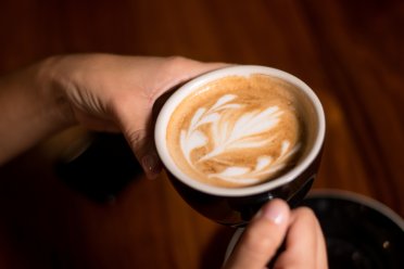 A person holding a black cup of coffee with latte art on top, over a wooden table.