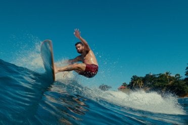 Man surfing a wave off the coast of Puerto Rico, carving across clear blue water with palm trees and shoreline in the background.