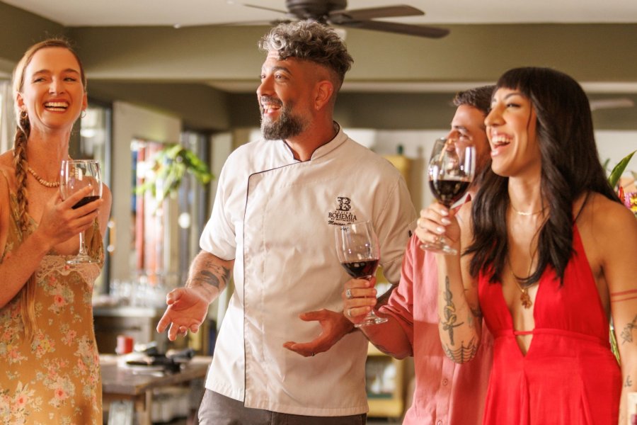 Chef leading a lively wine tasting indoors, as guests laugh and raise glasses around a table set with charcuterie and fresh ingredients.