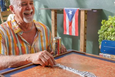 Older man laughing while playing dominoes at an outdoor table in Puerto Rico, with a Puerto Rican flag hanging in the background.
