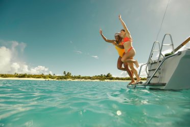 Two women in bright swimsuits jump joyfully from the back of a boat into clear turquoise ocean water near a tropical island shoreline in Puerto Rico under a sunny blue sky.