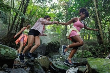 Three women hiking and carefully crossing rocks in a lush tropical rainforest in Puerto Rico, surrounded by dense greenery and mist.