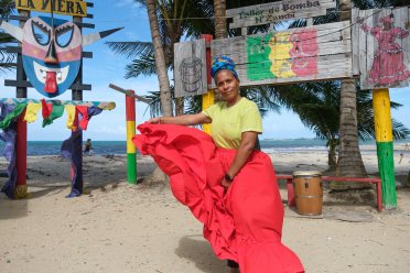 Sheila Osorio leading a traditional bomba dance workshop on the beach in Puerto Rico, wearing a vibrant red skirt with drums and cultural signs in the background.