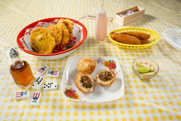 A colorful Puerto Rican fritura spread on a yellow checkered tablecloth, featuring alcapurrias, bacalaítos, rellenos de papa stuffed with ground beef, lime wedges, hot sauce, and domino tiles decorated with the Puerto Rico flag.