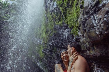 A couple embraces and smiles beneath a lush tropical waterfall, standing against moss-covered rocks as water cascades down beside them in a rainforest setting.