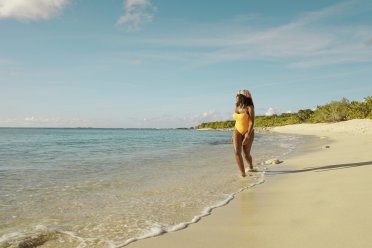 A woman in a yellow swimsuit walks along a sunny beach, letting gentle waves wash over her feet as turquoise water meets soft sand under a clear blue sky.