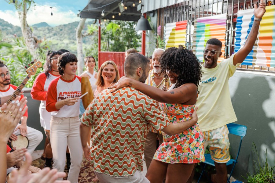 A joyful woman in a floral dress dances at an outdoor party in Cayey, Puerto Rico, accompanied by a traditional güiro player, colorful decor, and mountain views.