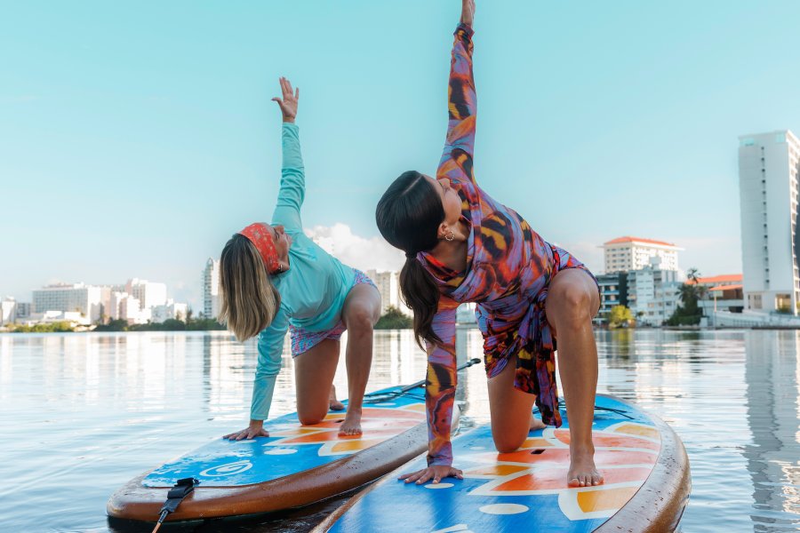 Two women practicing yoga on stand-up paddleboards in calm water with a city skyline in the background in Puerto Rico.