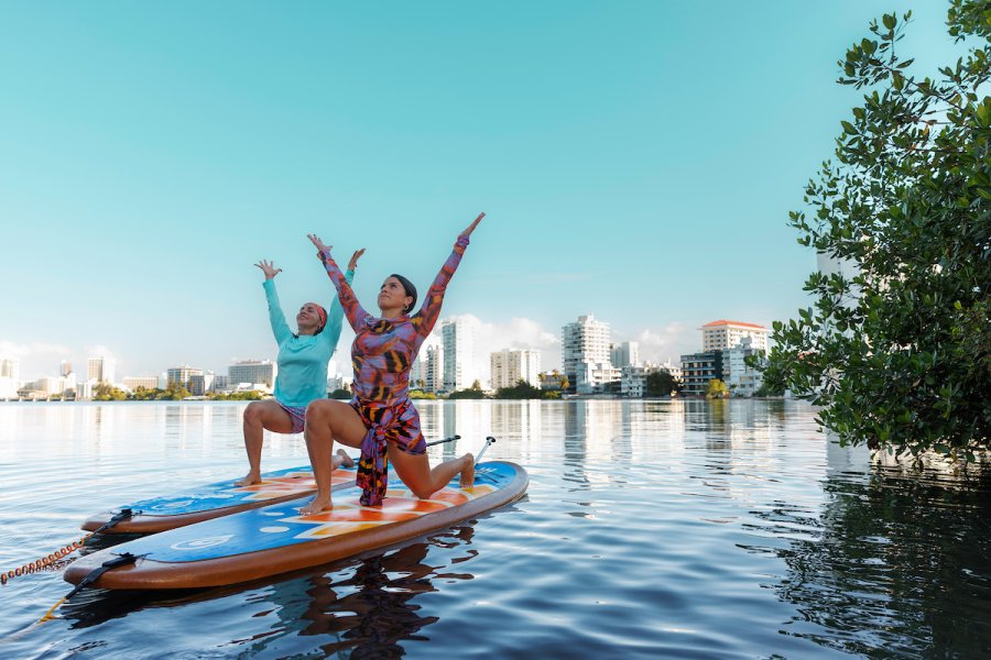 Two women practice paddleboard yoga on calm water in Puerto Rico, with a city skyline and mangroves in the background.