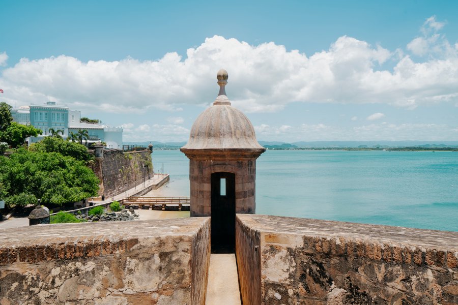 Historic stone fortification in Old San Juan overlooking the turquoise waters of San Juan Bay, with a sentry box, historic buildings, and mountains in the distance.