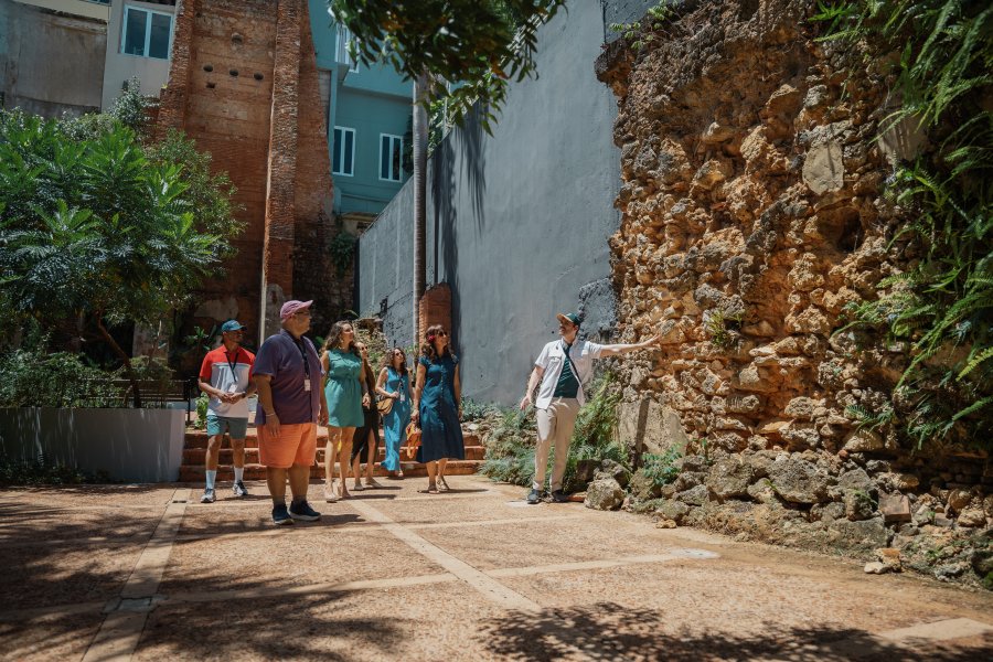 A tour guide points out historic stone ruins to a group visiting a courtyard in Old San Juan, Puerto Rico, surrounded by tropical greenery and colorful buildings.