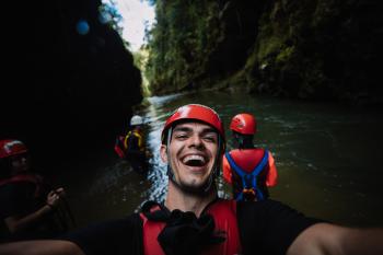 A guy in a helmet and life jacket standing in a river smiles at the camera 