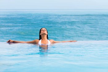 A woman relaxes with her head back in an infinity pool with the ocean in the background. Condado Vanderbilt Hotel, San Juan, Puerto Rico.