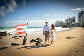 A bride and groom walk on the beach near the San Juan Marriott Resort & Stellaris Casino, with a Puerto Rican flag planted in the sand.