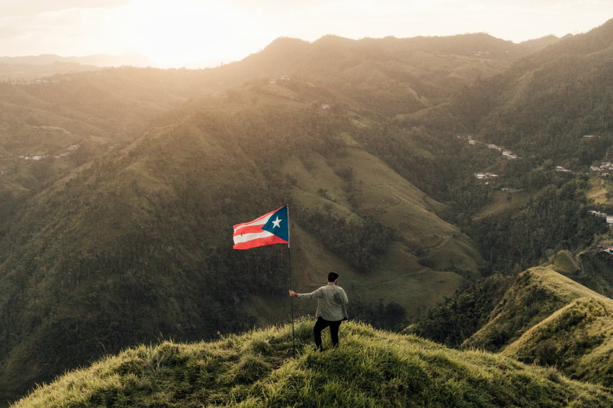 A man stands with a Puerto Rican flag atop a mountain summit as the sun sets.