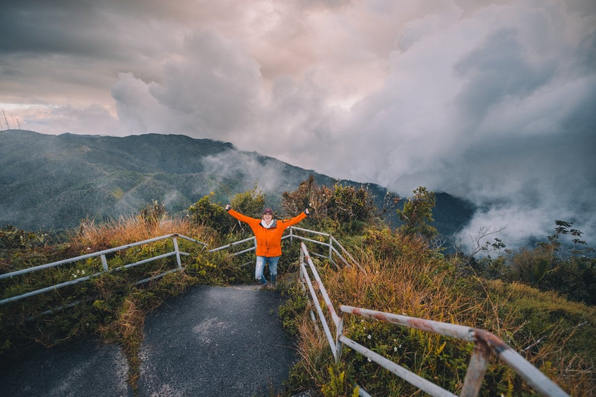 A woman raises her arms triumphantly after hiking up the side of a mountain.
