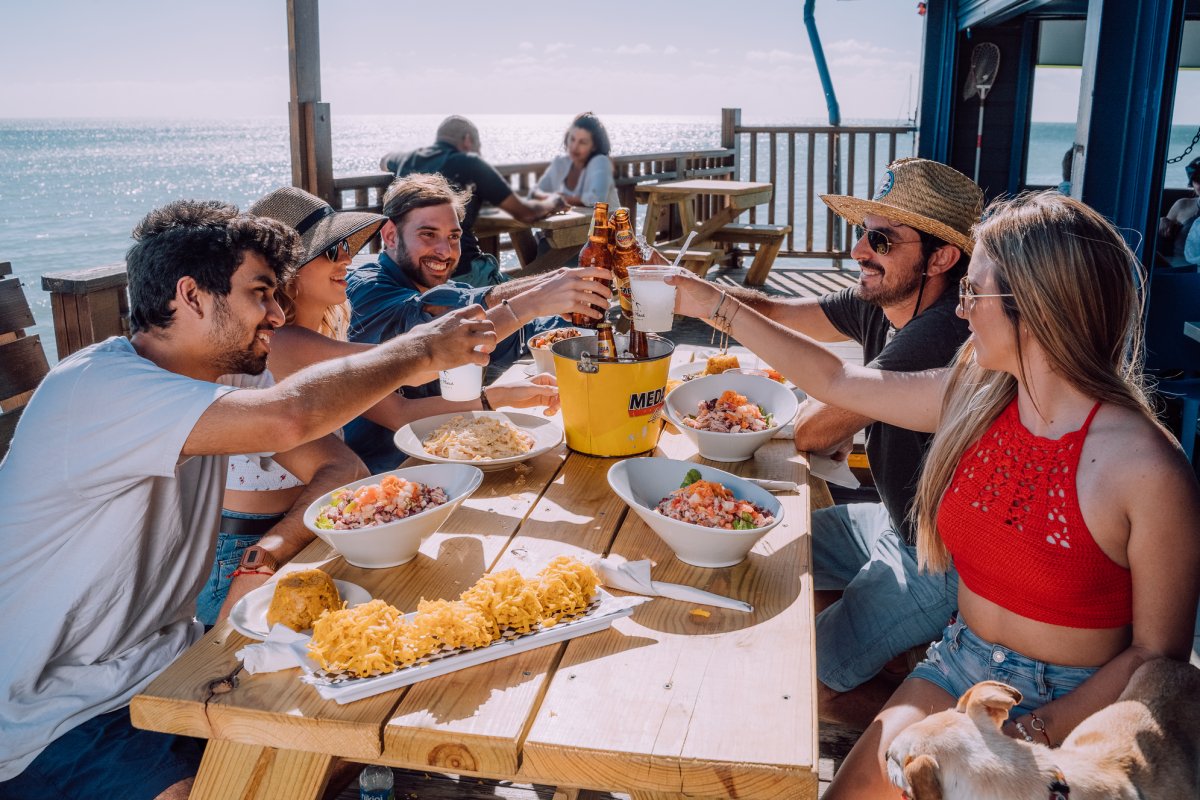A group of friends toasts at an outdoor table overlooking the sea.