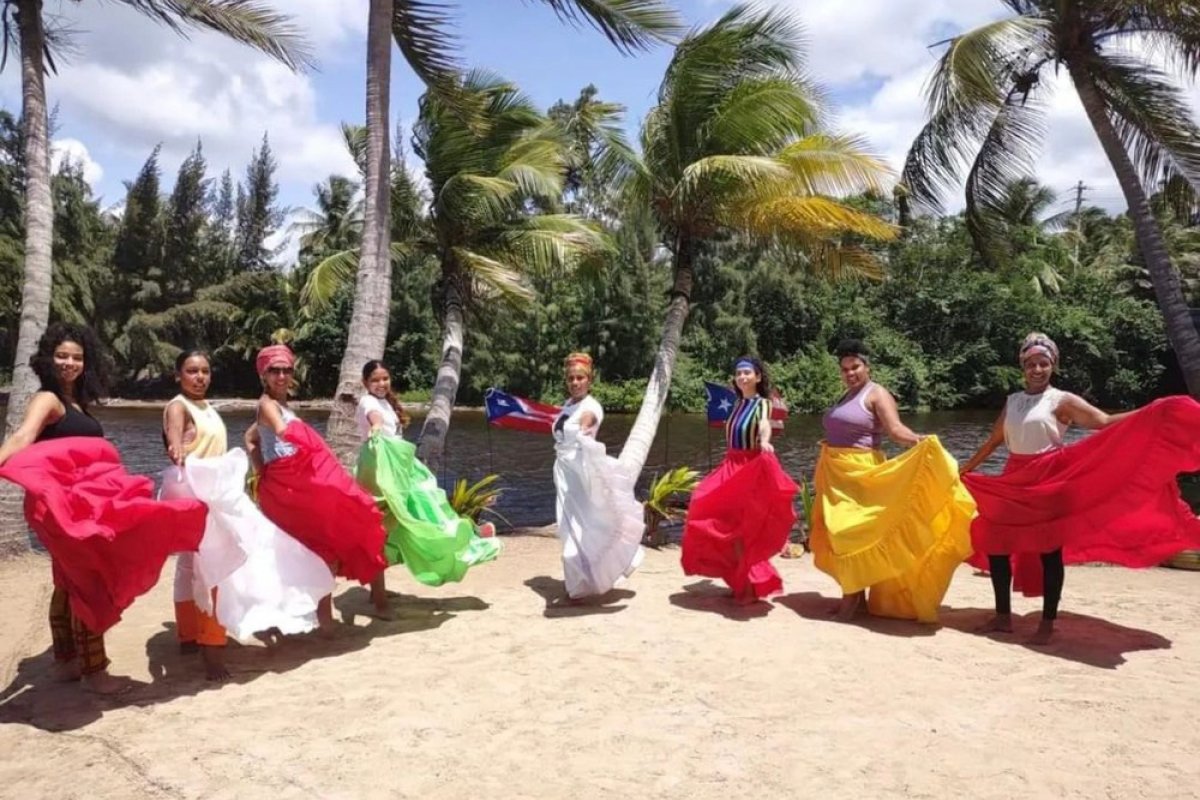 A group of performers in colorful traditional dresses stand in a semicircle smiling with palm trees behind them. Loiza, Puerto Rico.