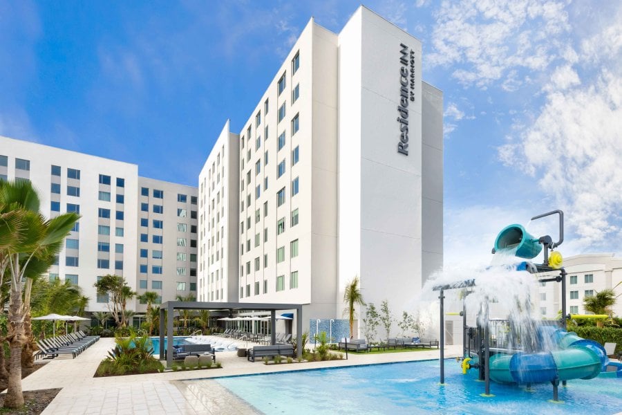Photo showing a swimming pool with water fountain play area, with a tall white hotel building in the background. Residence Inn by Marriott San Juan Isla Verde.