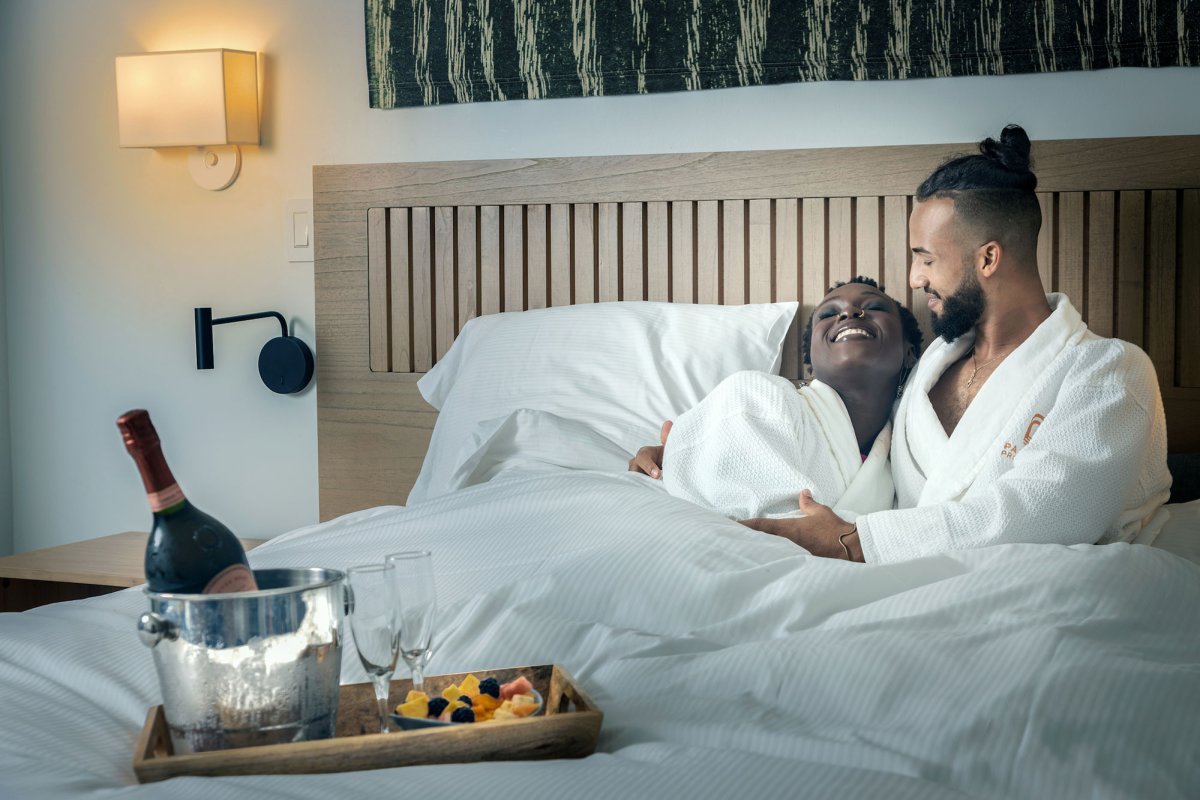 A couple lies in a hotel bed wearing white robes with a bottle of wine at the Hotel Palacio Provincial in San Juan, Puerto Rico.