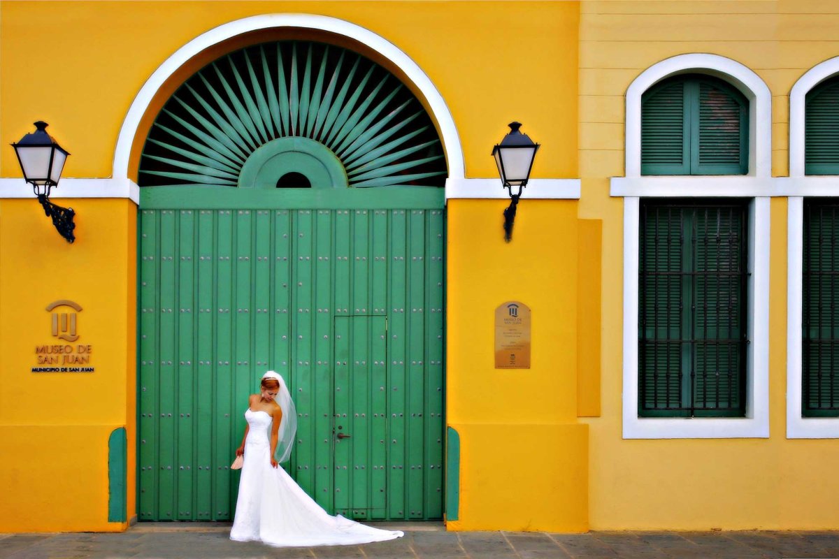 A bride poses in front of a yellow historic building with an arched teal door in San Juan, Puerto Rico.