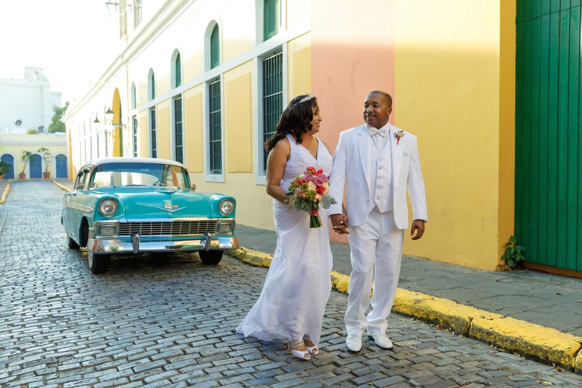 A wedding couple walks down a cobblestone street in Old San Juan with a teal-colored classic car parked behind them.