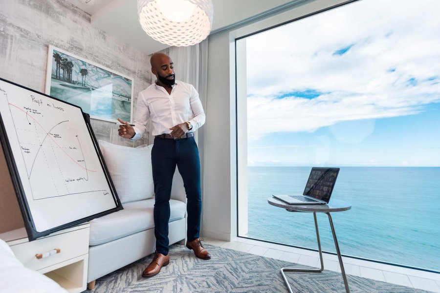 A man stands in front of a white board and a large window with an ocean view while making a remote presentation to his computer.