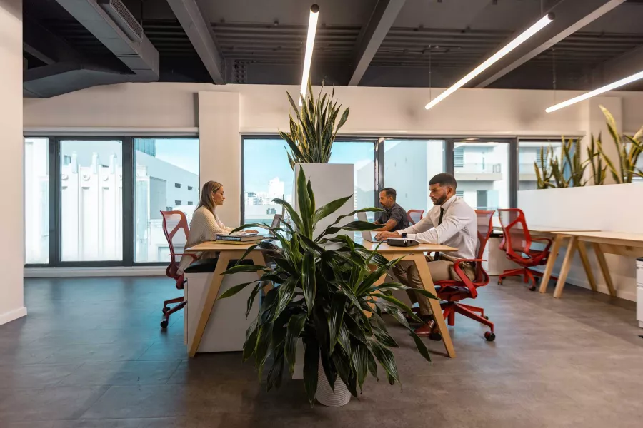 Three people work at a shared desk at a coworking space in San Juan, Puerto Rico, with a view of the city out of the windows.