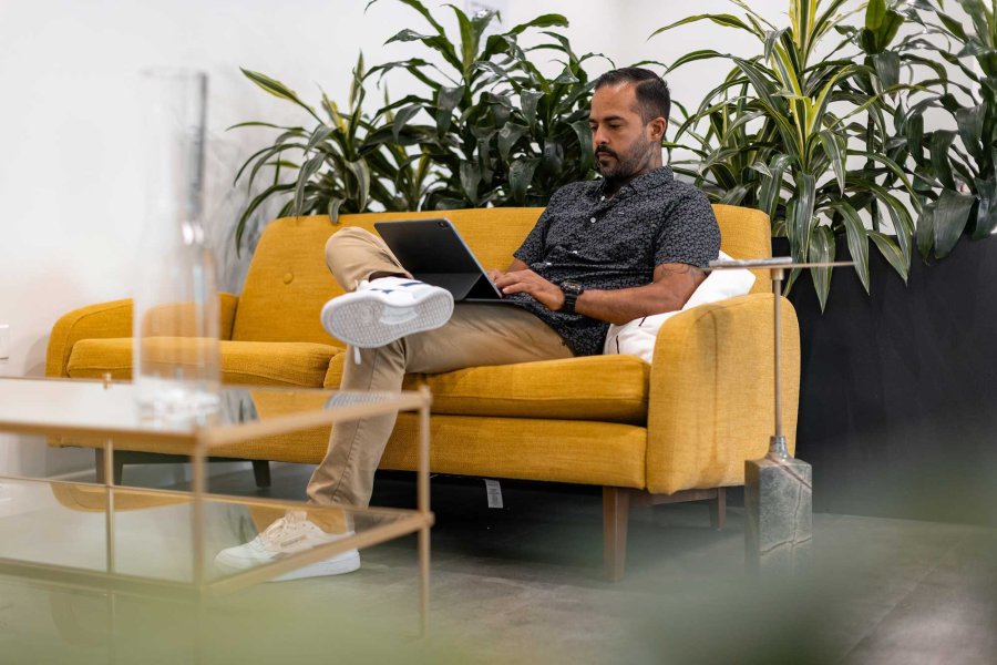 A man works on his laptop in a modern office while sitting on a yellow sofa.