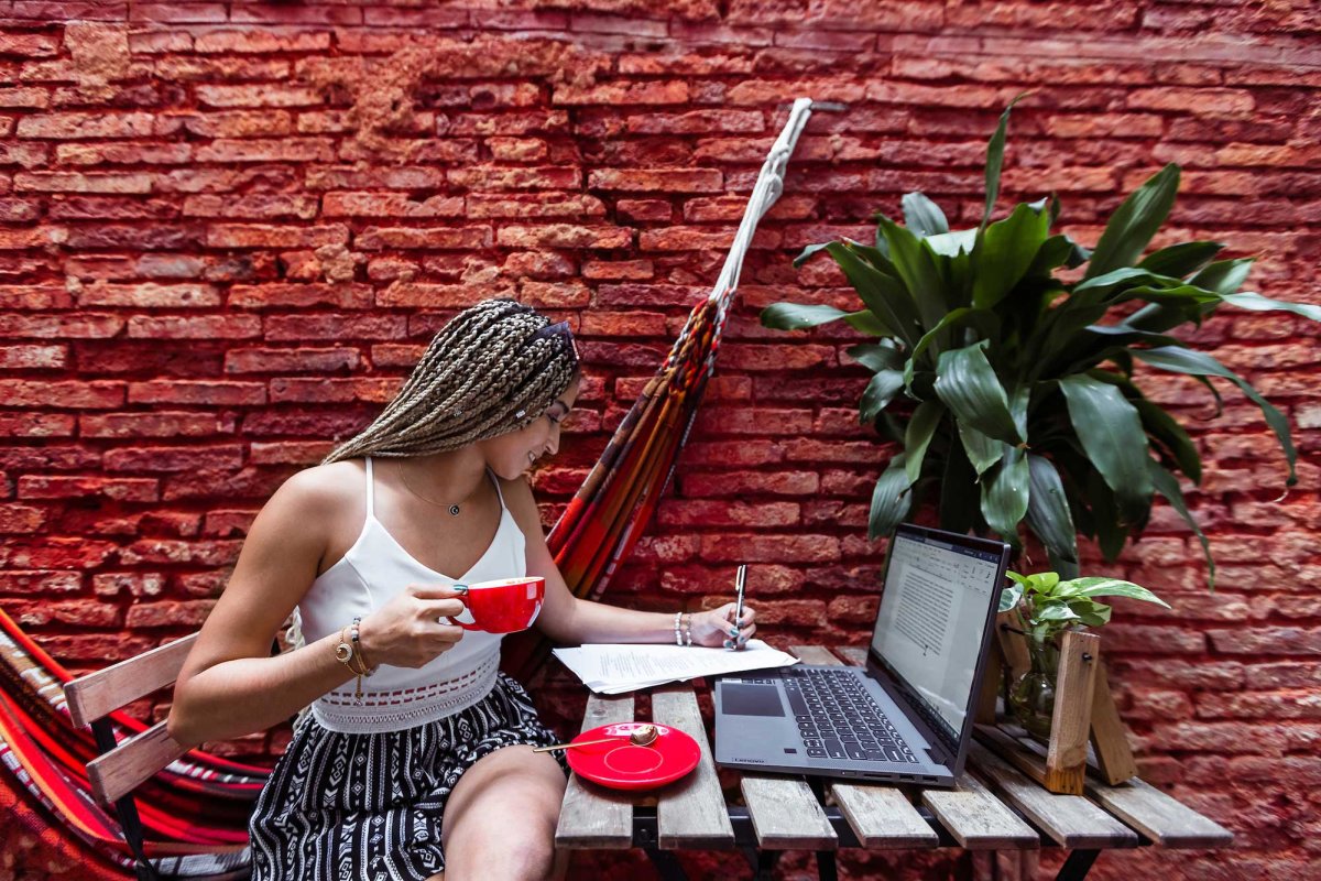 A woman works on her laptop computer at an outdoor desk against a vivid red brick wall.