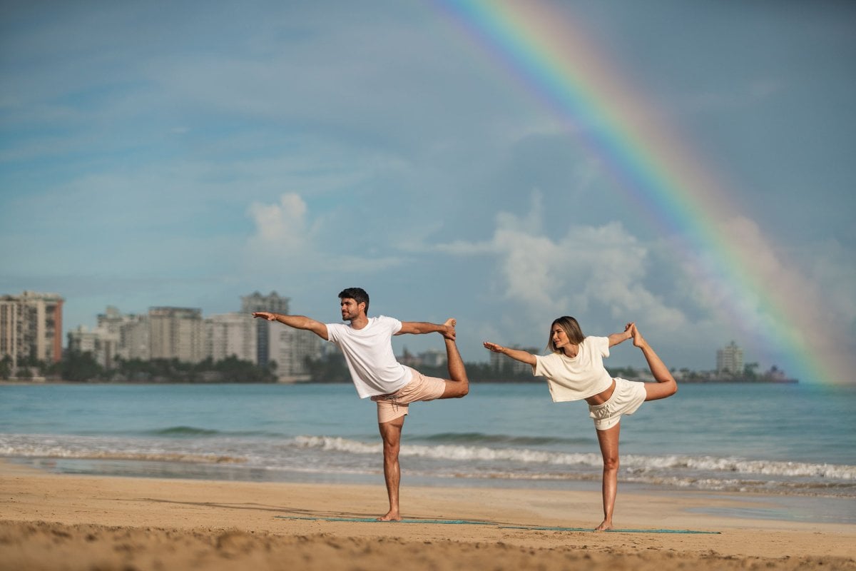 Una pareja practica yoga en una playa con un arco iris sobre sus cabezas en el hotel Fairmont El San Juan.