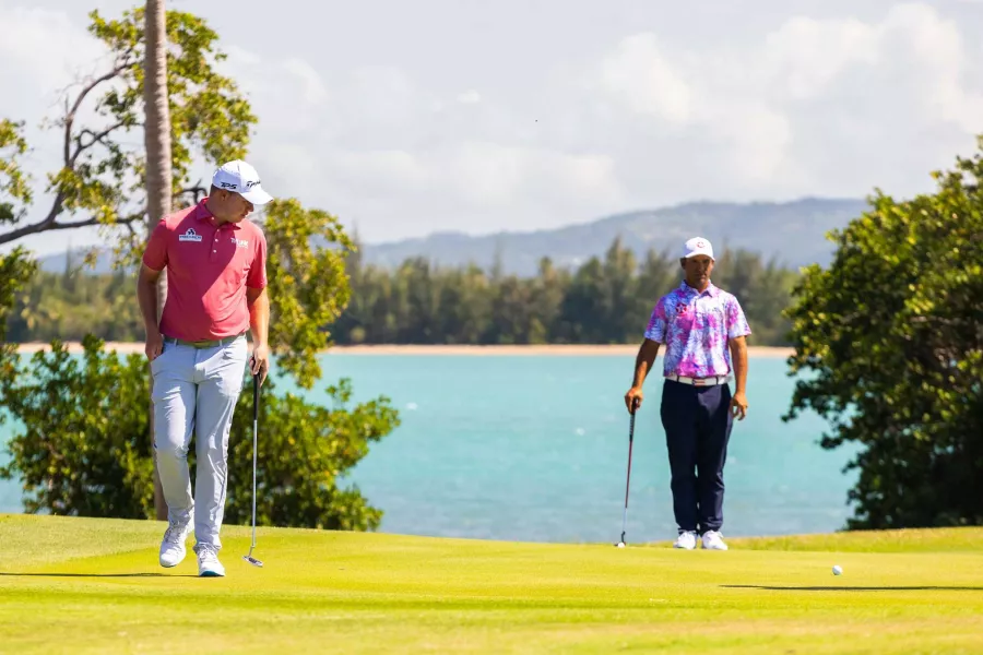 Dos golfistas están parados en el Club de Golf Grand Reserve en Río Grande, Puerto Rico, con agua azul y montañas en el fondo.