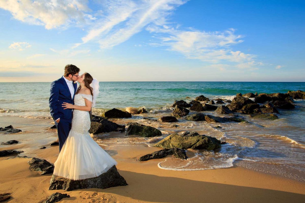 A newly wed bride and groom kiss while standing on a beach scattered with rocks in Puerto Rico. Photo by Noel Del Pilar.