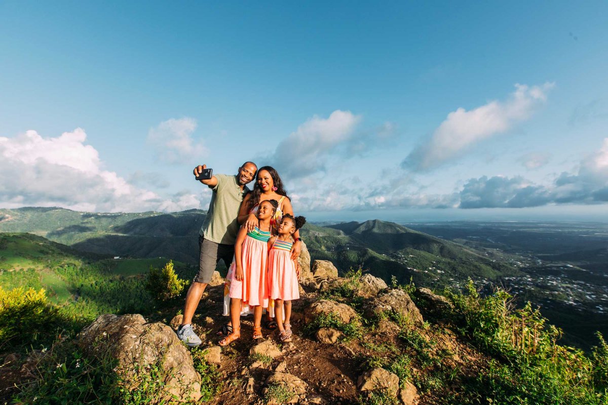 A mother, father, and two young girls pose for a selfie on top of a mountain with a dramatic backdrop in Puerto Rico.