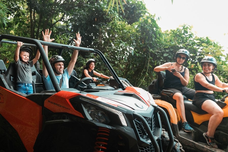 Man and son celebrating with their hands up inside an ATV going through the rainforest in Puerto Rico. The mother in another ATV and two other women on ATVs surround them.