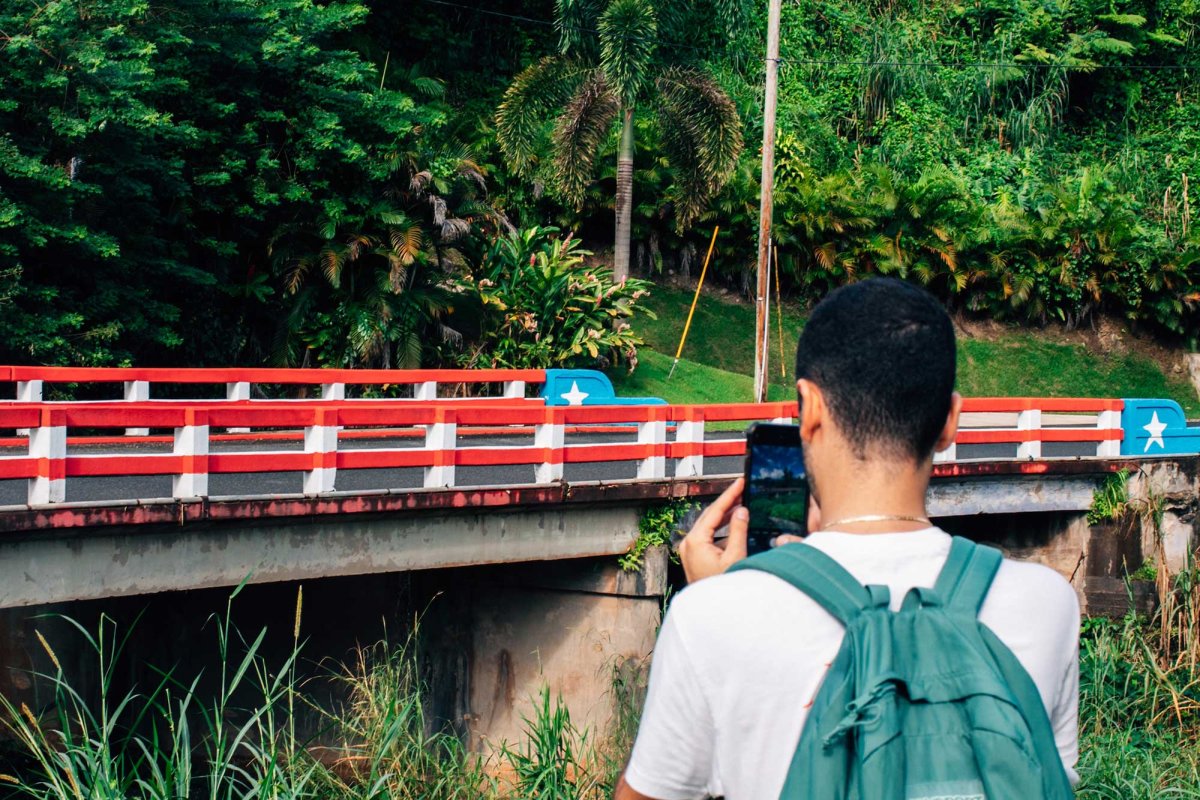 A man, seen from behind, takes a picture of a small bridge in Ciales, Puerto Rico, that is painted like the Puerto Rican flag.