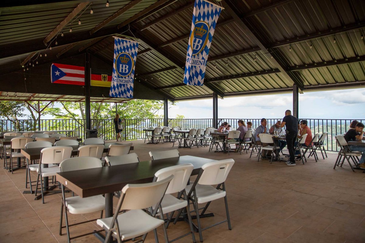 View of the open-air dining room of Casa Bavaria in Morovis, Puerto Rico, with German flags hanging from a pitched ceiling and expansive hillside views. 