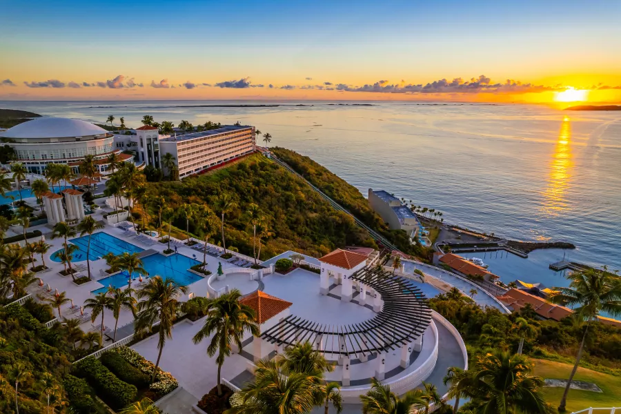 A view of El Conquistador Resort in Fajardo, Puerto Rico, overlooking the ocean at sunrise with pools, palm trees, and a marina.