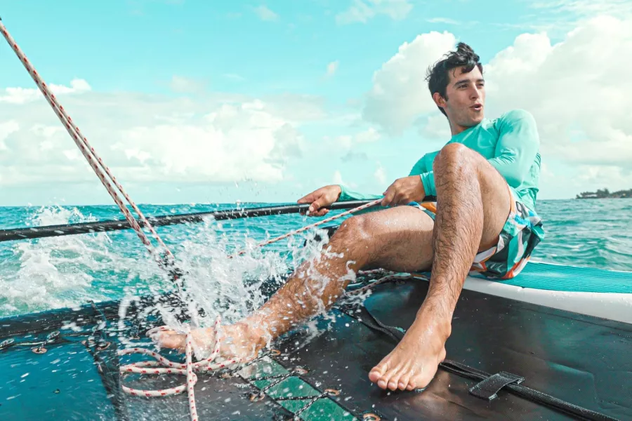 Man sailing a small catamaran in turquoise waters off Puerto Rico, pulling ropes as waves splash across the deck under a bright blue sky.