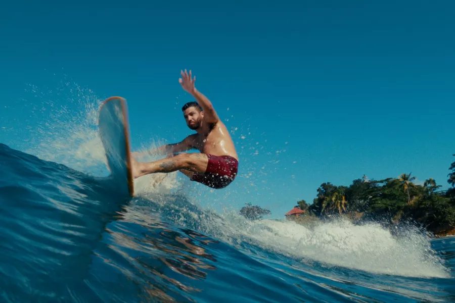 Man surfing a wave off the coast of Puerto Rico, carving across clear blue water with palm trees and shoreline in the background.