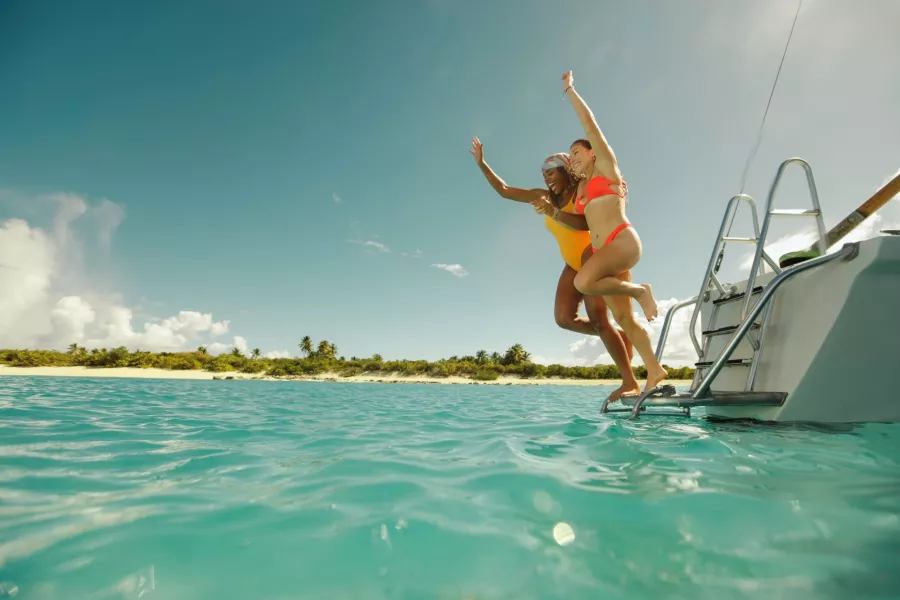 Two women in bright swimsuits jump joyfully from the back of a boat into clear turquoise ocean water near a tropical island shoreline in Puerto Rico under a sunny blue sky.