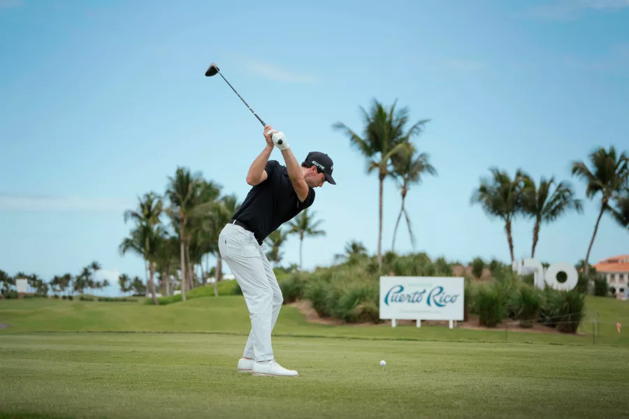 Golfer mid-swing on a lush Puerto Rico golf course with palm trees and a Puerto Rico sign in the background during the Puerto Rico Open.